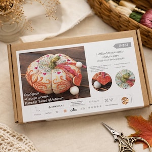 Pumpkin Heart of Autumn cross stitch kit packaging displayed on a soft neutral background. The presentation box features the finished pincushion design and highlights premium materials including Zweigart Belfast linen, hand-dyed DMC floss, decorative stitches, ceramic components, and complete stitching instructions.