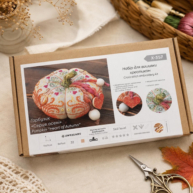 Pumpkin Heart of Autumn cross stitch kit packaging displayed on a soft neutral background. The presentation box features the finished pincushion design and highlights premium materials including Zweigart Belfast linen, hand-dyed DMC floss, decorative stitches, ceramic components, and complete stitching instructions.
