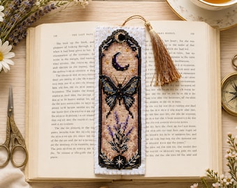 May include: A rectangular cross-stitch bookmark featuring a dark butterfly, a crescent moon, and lavender sprigs on a beige fabric background. The bookmark is bordered in black, topped with a gold tassel, and rests on an open book page.