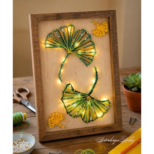 Peut inclure: Une œuvre d'art encadrée en fil de fer représentant deux feuilles de ginkgo de tons verts et jaunes, illuminées par de petites lumières. L'œuvre est placée sur un fond en bois clair dans un cadre marron rustique. Des accents supplémentaires en fil jaune sont visibles.