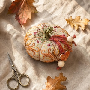 Pumpkin pincushion sampler photographed on soft linen fabric with autumn leaves and vintage embroidery scissors. The composition highlights the decorative stitching, ceramic heart charm, hand-dyed thread, and warm seasonal palette designed for embroidery enthusiasts.