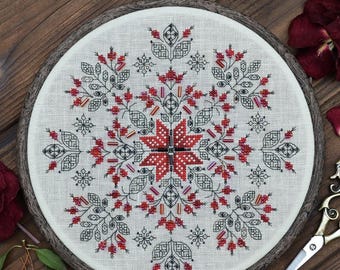 以下が含まれることがあります： 円形の刺繍枠に収められたオフホワイトの布地に、左右対称のクロスステッチ模様が施されています。中央の赤い星のモチーフを囲むように、黒い幾何学的な花模様と小さな赤いガラスビーズが配置されています。