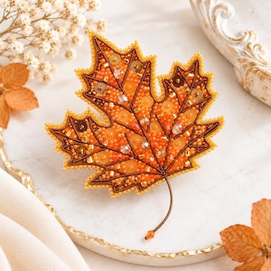 Peut inclure: Une broche en forme de feuille d'érable ornée de perles de rocaille, de sequins et de cristaux orange et marron. La feuille présente un contour perlé détaillé et une tige fine et incurvée, mettant en valeur une broderie complexe.