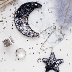 May include: A crescent moon brooch and a star brooch connected by a silver-toned metal chain. Both pieces are heavily embellished with dark grey, black, and clear beads, crystals, and pearls. Two silver-toned metal rings are positioned nearby.