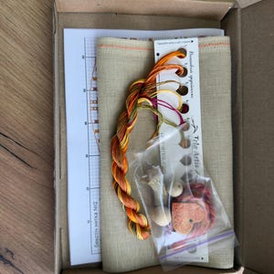 May include: A cross-stitch kit in a cardboard box containing a piece of beige fabric, a printed pattern chart, a thread organizer card with numbered holes, multi-colored embroidery floss, a small plastic bag with wooden beads, and a heart-shaped charm.
