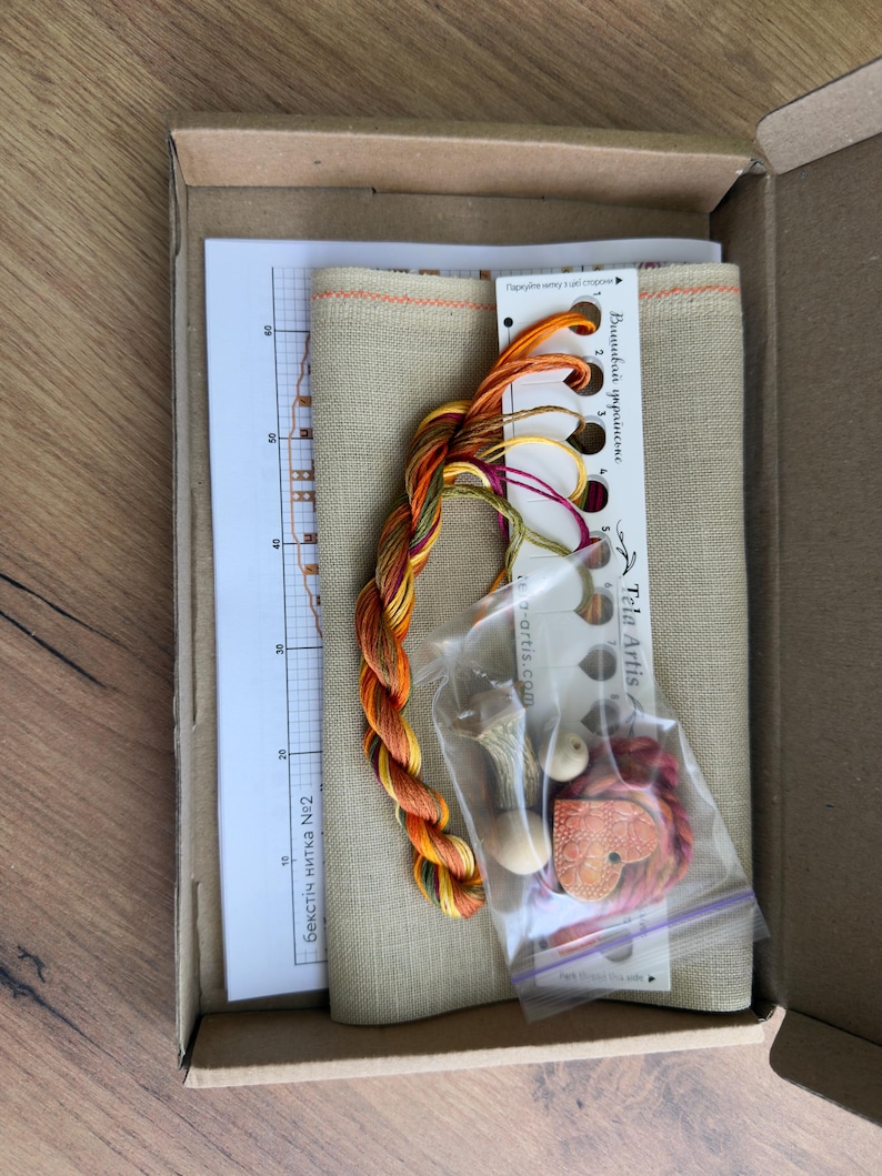 May include: A cross-stitch kit in a cardboard box containing a piece of beige fabric, a printed pattern chart, a thread organizer card with numbered holes, multi-colored embroidery floss, a small plastic bag with wooden beads, and a heart-shaped charm.