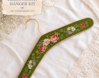 May include: A cross stitch hanger kit featuring a green fabric-covered clothes hanger with a gold metal hook. The hanger is embroidered with a floral pattern of pink, red, white, and purple flowers. The text reads CROSS STITCH HANGER KIT.