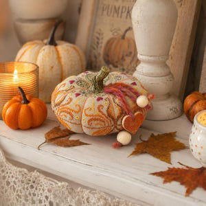 Autumn pumpkin pincushion displayed as seasonal home décor among candles, pumpkins, and fall leaves. The embroidered pincushion combines counted cross stitch, decorative stitches, ceramic accents, and hand-dyed floss to create a charming autumn display piece.