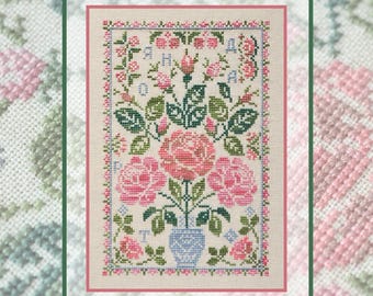May include: A cross-stitch embroidery on light beige fabric featuring a central blue vase filled with pink roses and green leaves. The design is framed by a decorative border of small pink and blue flowers, with the letters A, H, A, and O integrated.