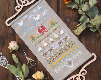 May include: A rectangular linen fabric wall hanging featuring a detailed cross-stitch pattern of a red house, white geese, yellow ducklings, and floral borders. The fabric is held by two light-coloured wooden scroll hangers at the top and bottom.