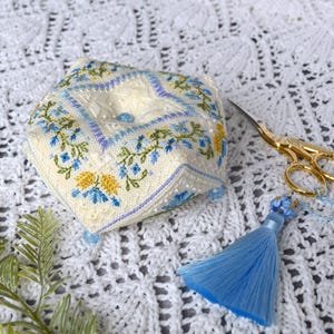 May include: A square, cream-colored fabric pincushion featuring intricate embroidery of blue and yellow floral patterns, accented with light blue beads at the corners and center. A pair of gold-toned metal embroidery scissors with a blue tassel.