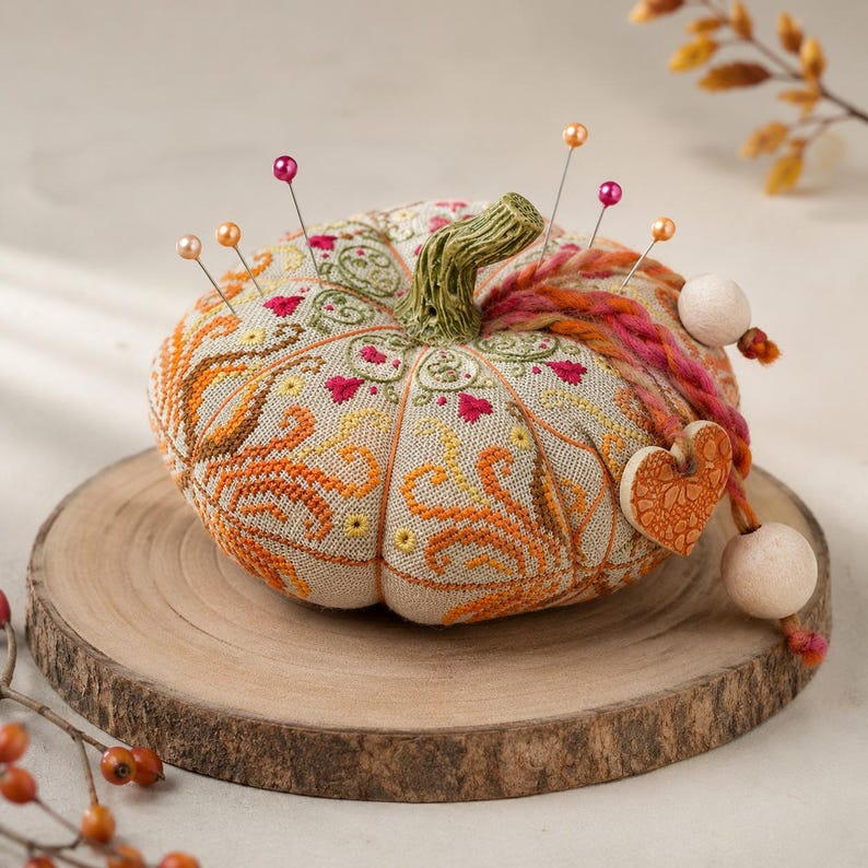 Autumn pumpkin pincushion cross stitch sampler finished as a decorative three-dimensional pumpkin. The handmade pincushion features intricate counted cross stitch, decorative stitches, hand-dyed DMC floss, a ceramic pumpkin stem, ceramic heart charm, wooden beads, and autumn-inspired colors. Displayed on a rustic wooden surface with seasonal accents.