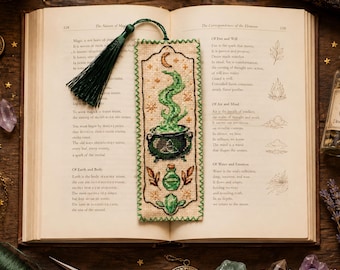 May include: A rectangular cross-stitch bookmark on beige fabric featuring a black cauldron with green swirling smoke, a small green potion bottle, and a green crystal. The bookmark has a dark green tassel and a decorative green stitched border.
