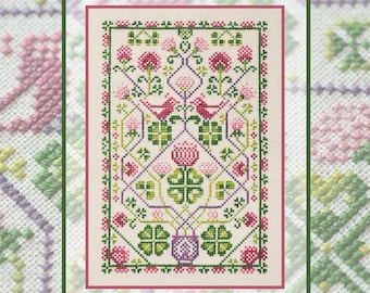 May include: A cross-stitch embroidery pattern on cream fabric featuring a symmetrical design of pink flowers, green clover leaves, and two small birds facing each other. The rectangular composition is enclosed by a decorative green border.