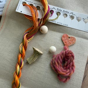 May include: A beige linen fabric base with a white thread organizer labeled Tela Artis, featuring a braided strand of variegated orange, yellow, and purple embroidery floss, two round wooden beads, a small mushroom-shaped charm, and a pink heart charm.