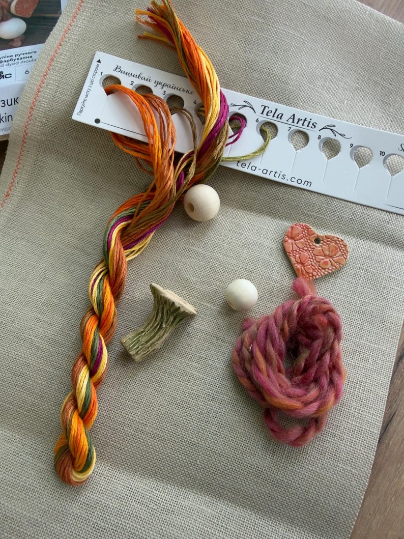 May include: A beige linen fabric base with a white thread organizer labeled Tela Artis, featuring a braided strand of variegated orange, yellow, and purple embroidery floss, two round wooden beads, a small mushroom-shaped charm, and a pink heart charm.