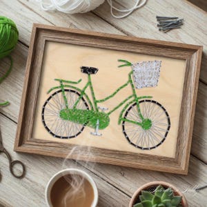May include: A framed artwork featuring a green bicycle crafted with string on a light wood background. The bicycle has a white basket and black tires. The frame is a warm brown color. A cup of coffee and a small succulent plant are in the foreground.