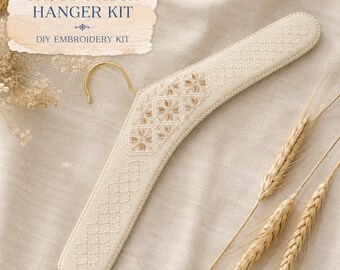May include: A beige fabric-covered clothes hanger featuring intricate cross-stitch embroidery in light brown and white thread. The hanger has a gold-toned metal hook and includes a label that reads CROSS STITCH HANGER KIT DIY EMBROIDERY KIT.