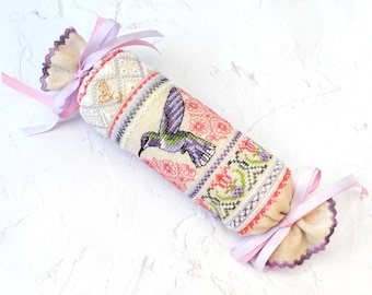 May include: A cylindrical fabric pincushion featuring cross-stitch embroidery of a hummingbird and floral patterns in shades of green, purple, and pink. The ends are gathered and tied with light pink ribbons, and a small gold hummingbird charm is attached.