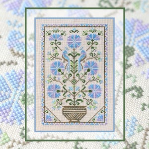 May include: A cross-stitch embroidery pattern on light beige fabric featuring a central brown basket holding a symmetrical floral arrangement with blue and purple flowers, green stems, and two blue birds facing each other in a rectangular border.
