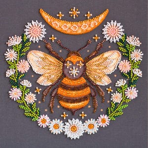 Könnte beinhalten: Eine Perlen-Stickerei mit einer Biene mit ausgebreiteten Flügeln, umgeben von einem Kranz aus weißen und rosa Blumen und einem Halbmond mit Sternen darüber.