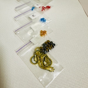 Puede incluir: Variedad de cuentas en peque&ntilde;as bolsas transparentes con cierre de cremallera. Las cuentas son de varios colores, incluyendo rojo, azul, dorado y negro. Algunas bolsas tienen peque&ntilde;as etiquetas blancas con n&uacute;meros. Las cuentas est&aacute;n sobre una superficie blanca texturizada.