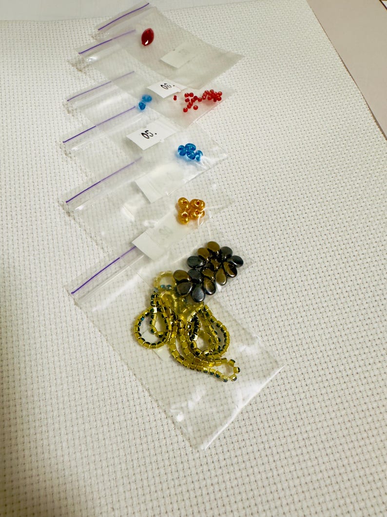 Puede incluir: Variedad de cuentas en peque&ntilde;as bolsas transparentes con cierre de cremallera. Las cuentas son de varios colores, incluyendo rojo, azul, dorado y negro. Algunas bolsas tienen peque&ntilde;as etiquetas blancas con n&uacute;meros. Las cuentas est&aacute;n sobre una superficie blanca texturizada.