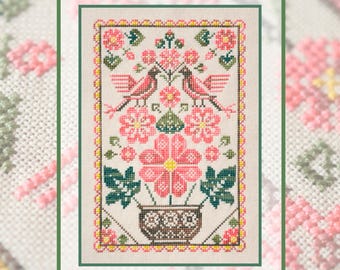 May include: A cross-stitch embroidery on light fabric featuring two birds perched above a central flower in a decorative pot. The design includes pink, green, and brown thread colours, surrounded by a yellow and pink geometric border pattern.