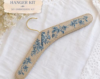 May include: A beige fabric-covered clothes hanger featuring a blue cross-stitch pattern of a bird and floral motifs. The hanger includes a gold-toned metal hook. The text "CROSS STITCH HANGER KIT" and "DIY EMBROIDERY KIT" is visible above the hanger.