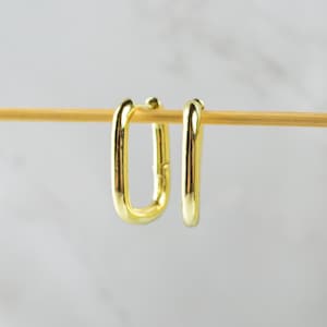 May include: A pair of gold-toned hoop earrings with a rectangular shape. The earrings are hanging from a wooden dowel.