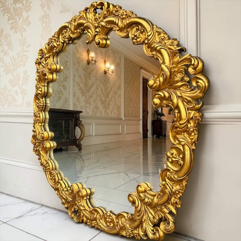 May include: Ornate, oval-shaped gold mirror with elaborate, swirling details. The mirror is leaning against a white wall, reflecting a room with patterned wallpaper and a marble floor. The frame is a rich, metallic gold.