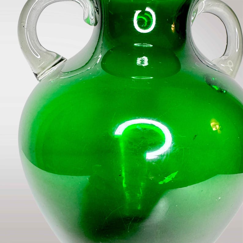 May include: A green glass vase with two handles. The vase has a rounded body and a smooth, glossy finish.