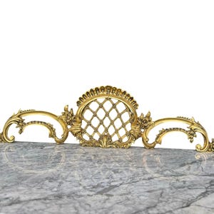 May include: Ornate gold-colored decorative architectural element. The design features a central oval with a diamond pattern, flanked by scrolling floral accents. The piece is likely made of metal.