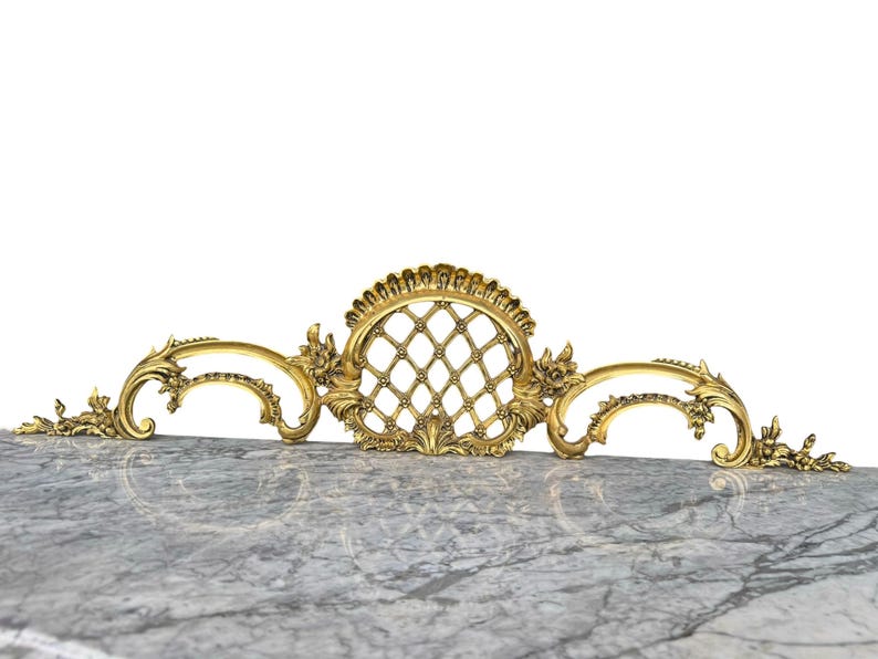 May include: Ornate gold-colored decorative architectural element. The design features a central oval with a diamond pattern, flanked by scrolling floral accents. The piece is likely made of metal.