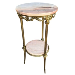May include: An antique side table with a round, pink and white marble top. The table has a second, smaller marble shelf and is supported by three brass legs. The brass frame features ornate floral detailing. The table is approximately 76 cm tall.