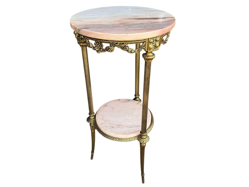 May include: An antique side table with a round, pink and white marble top. The table has a second, smaller marble shelf and is supported by three brass legs. The brass frame features ornate floral detailing. The table is approximately 76 cm tall.