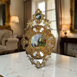 May include: An ornate, gold-coloured wall decoration featuring a central circular painting of figures in a landscape. The frame has intricate floral and scrollwork designs. A smaller circular painting is at the top. The decor is displayed on a white marble surface.