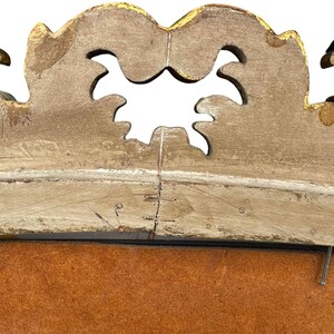 May include: Close-up of a decorative, ornate, antique wooden frame. The frame is painted in a distressed, off-white color with gold accents. The top of the frame has a scalloped design with cut-out shapes. The frame appears to be aged.