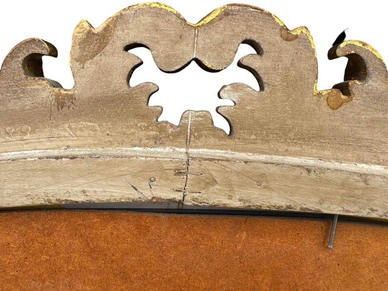 May include: Close-up of a decorative, ornate, antique wooden frame. The frame is painted in a distressed, off-white color with gold accents. The top of the frame has a scalloped design with cut-out shapes. The frame appears to be aged.