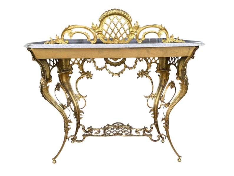 May include: An antique gold-colored console table with ornate detailing. The table features a marble top and intricate, curved legs. The design includes floral and scrollwork accents, with a decorative back panel. The table's height is approximately 30 inches.