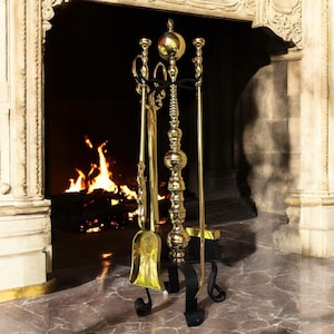 May include: A brass fireplace tool set including a shovel, poker, brush, and stand. The tools feature intricate detailing and are positioned in front of a fireplace with a burning fire. The stand is black with curved feet.