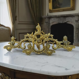 May include: Ornate gold-colored decorative metal piece with floral and scrollwork designs. The symmetrical piece features a central flower and is likely a decorative element for furniture or a wall.