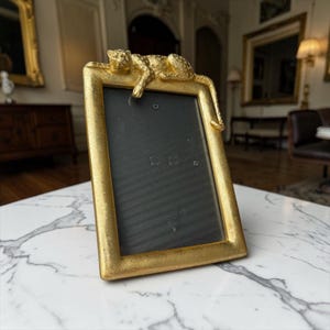 Op de afbeelding: Een goudkleurige rechthoekige fotolijst met een decoratieve luipaard die op de bovenrand rust. De lijst heeft een zwart interieur en wordt tentoongesteld op een wit marmeren oppervlak. De lijst is een decoratief woondecoratie-item.
