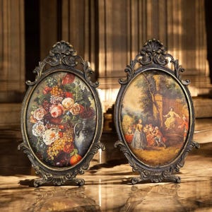 May include: Two antique oval picture frames with ornate bronze-colored metalwork. One frame features a still life of flowers, the other a pastoral scene with figures. Both are displayed on a marble surface.