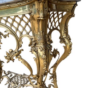 May include: Ornate gold-colored console table with a marble top. The table features intricate floral and lattice detailing, with curved legs and decorative accents. The design is reminiscent of antique furniture.
