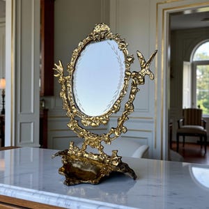 Puede incluir: Un espejo de tocador dorado y ornamentado, con un espejo ovalado. El marco presenta un diseño floral detallado. La base tiene forma de concha decorativa. El espejo está sobre una superficie de mármol. Altura aproximada del espejo: 30 cm.