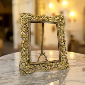 Puede incluir: Marco de fotos dorado y ornamentado con un centro vacío, sostenido por un pequeño caballete. El marco presenta intrincados adornos y florituras decorativas. El marco se muestra sobre una superficie de mármol.