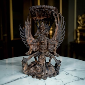 Puede incluir: Una escultura de madera de color marrón oscuro, tallada a mano con intrincados detalles, que representa una figura alada montada sobre una criatura. La talla muestra plumas detalladas, expresiones faciales y una parte superior en forma de cuenco.