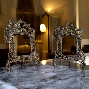 Può includere: Due cornici antiche in bronzo con dettagli ornati. Ogni cornice presenta un'apertura rettangolare, un bordo decorativo e una scena scolpita di una persona a cavallo. Le cornici sono esposte su una superficie di marmo.