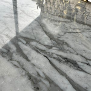 May include: Close-up of a white marble surface with gray veining. The polished stone reflects light, creating a glossy appearance. The surface is part of a larger piece, possibly a table or countertop.
